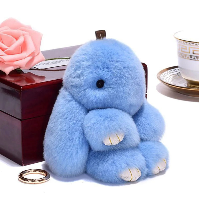 A fluffy light blue plush bunny keychain resting on a dark wooden box next to a pink rose.