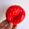 A hand holds a bright red strawberry squishy toy with green leaves against a white background.