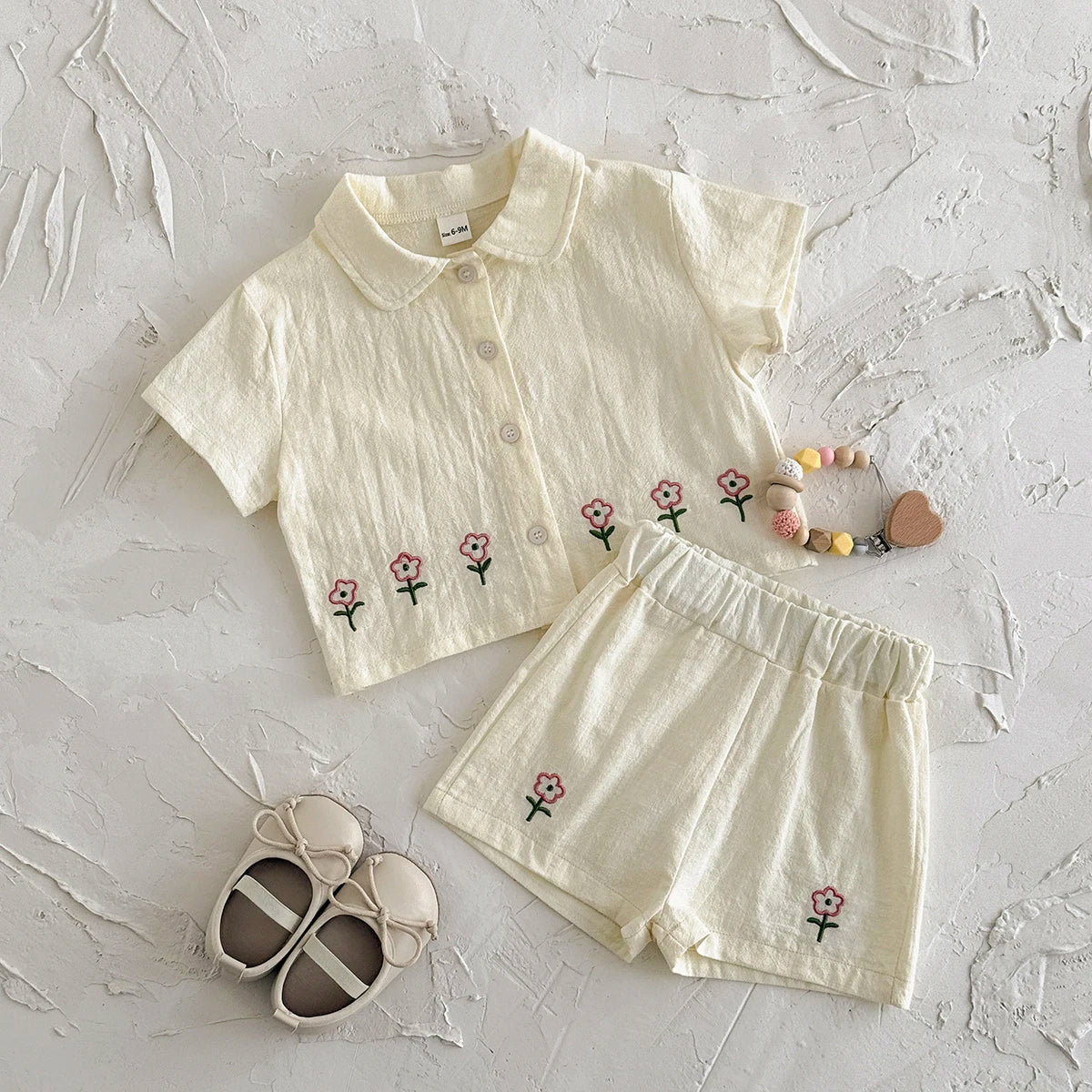 A cream baby cotton shirt and shorts set with a delicate pink flower pattern laid out on a white surface.