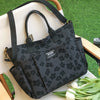 A black canvas tote bag with multiple pockets is displayed on a wooden surface against a green background.