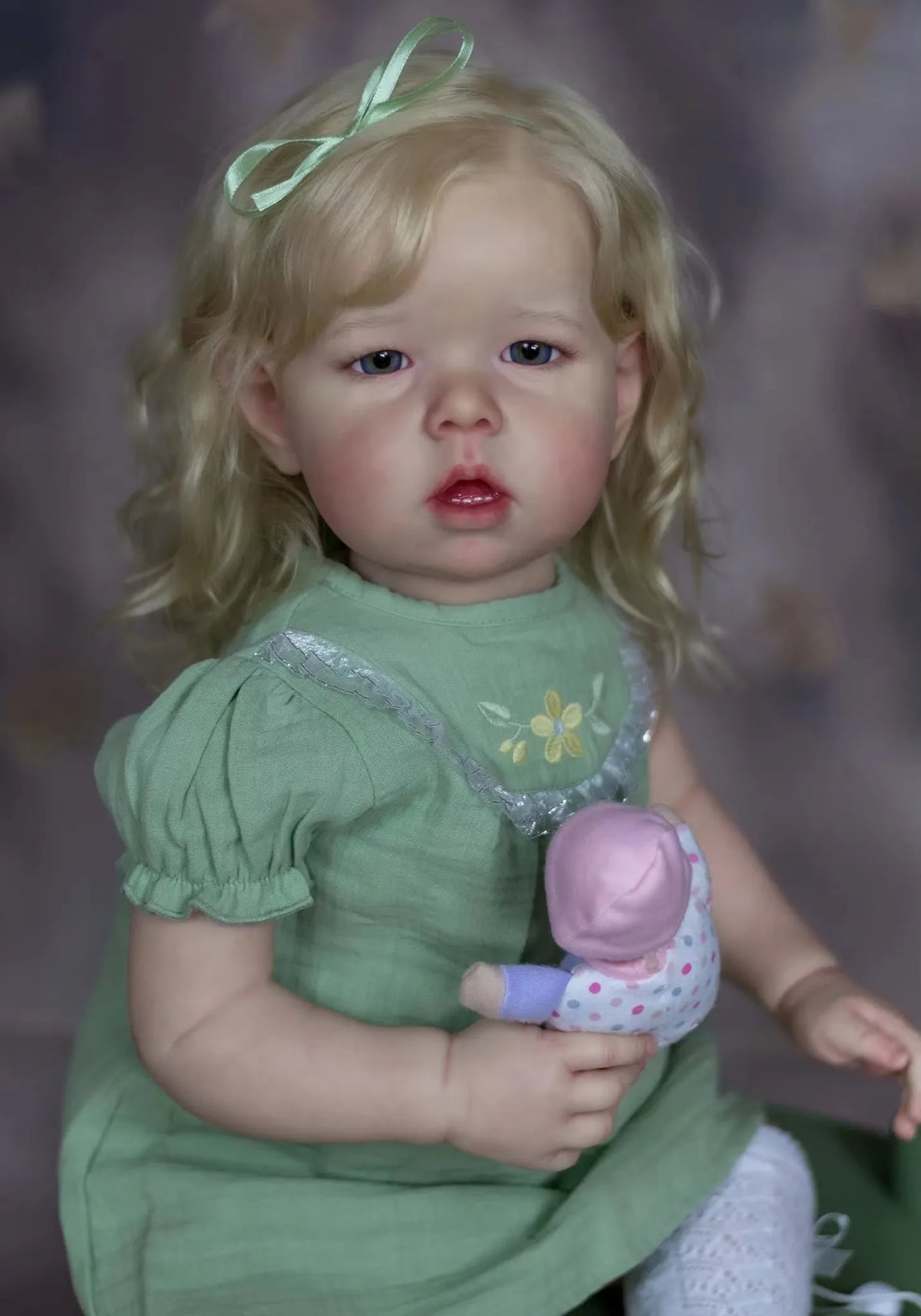 A realistic reborn baby doll with soft cloth body, featuring blonde hair and a green dress.