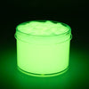 A clear jar of glow-in-the-dark crystal slime with a bright green luminescence and a layer of foamy substance on top.