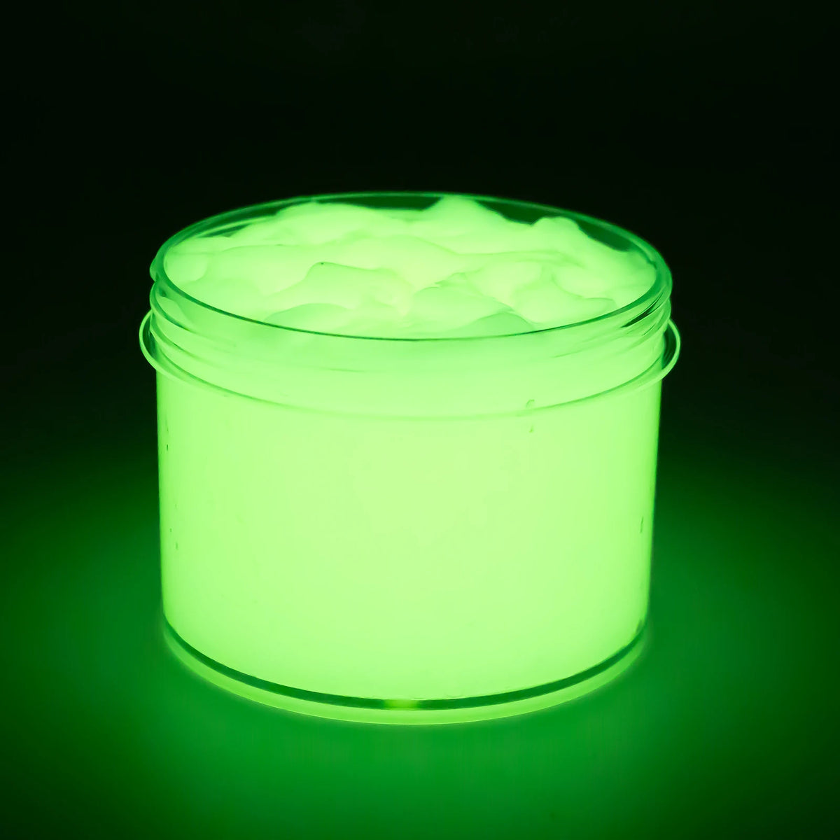 A clear jar of glow-in-the-dark crystal slime with a bright green luminescence and a layer of foamy substance on top.