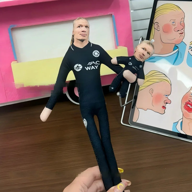A male football star stuffed doll wearing a black jersey is held by a hand in front of a pink display box.
