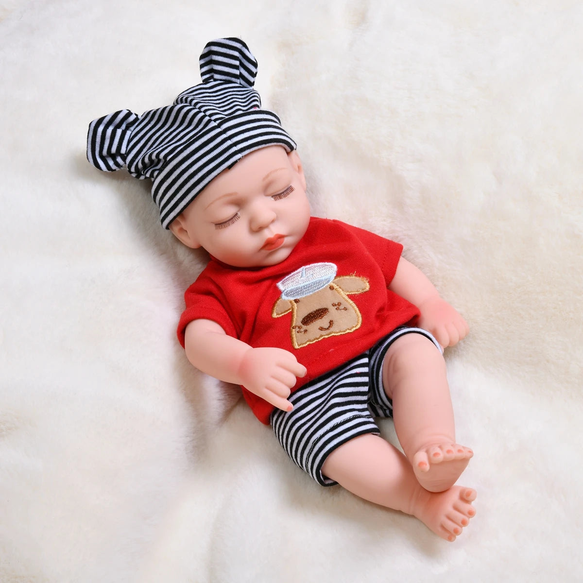 A lifelike vinyl reborn doll with a striped hat and red shirt lying on a white fluffy surface.
