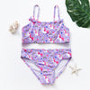 A purple girls two-piece swimsuit set featuring white unicorns and rainbow patterns, laid on a white surface.