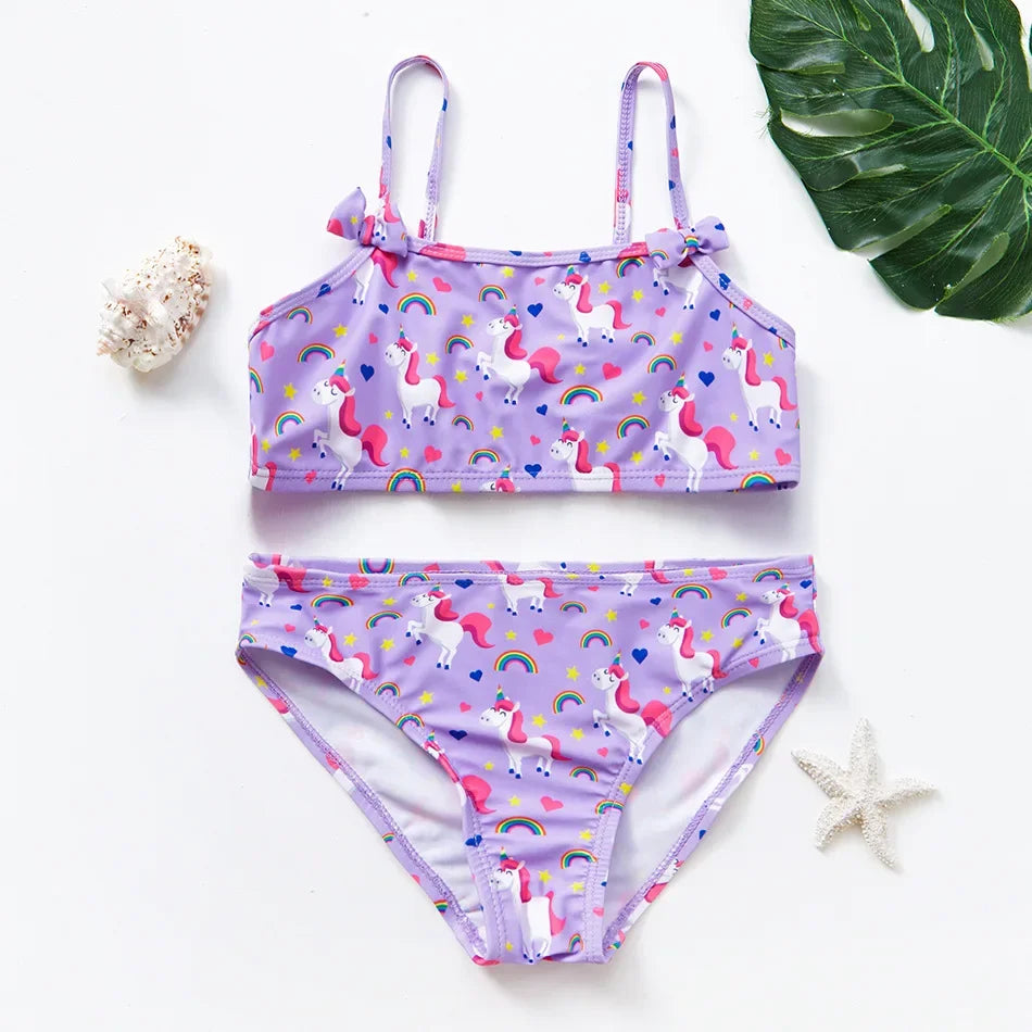 A purple girls two-piece swimsuit set featuring white unicorns and rainbow patterns, laid on a white surface.