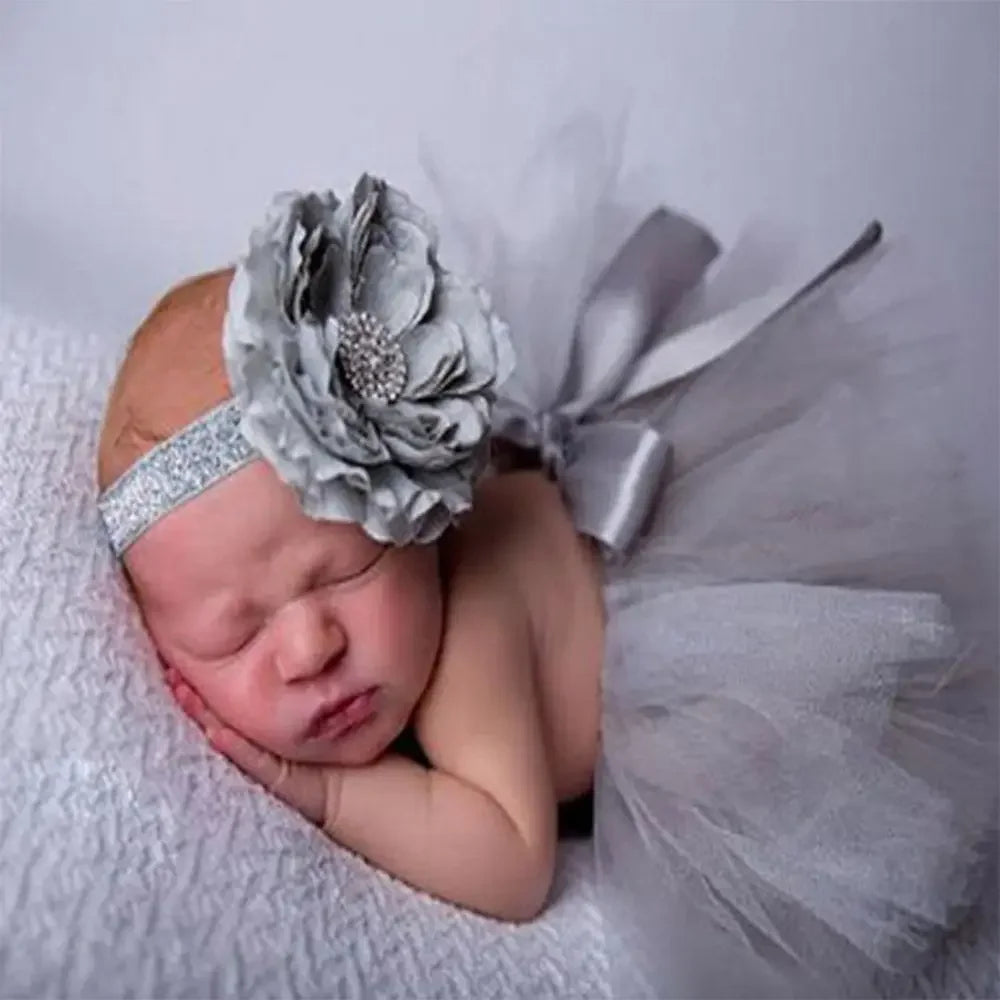 A newborn baby girl sleeping peacefully wearing a silver crown headband and surrounded by sheer gray tulle fabric.