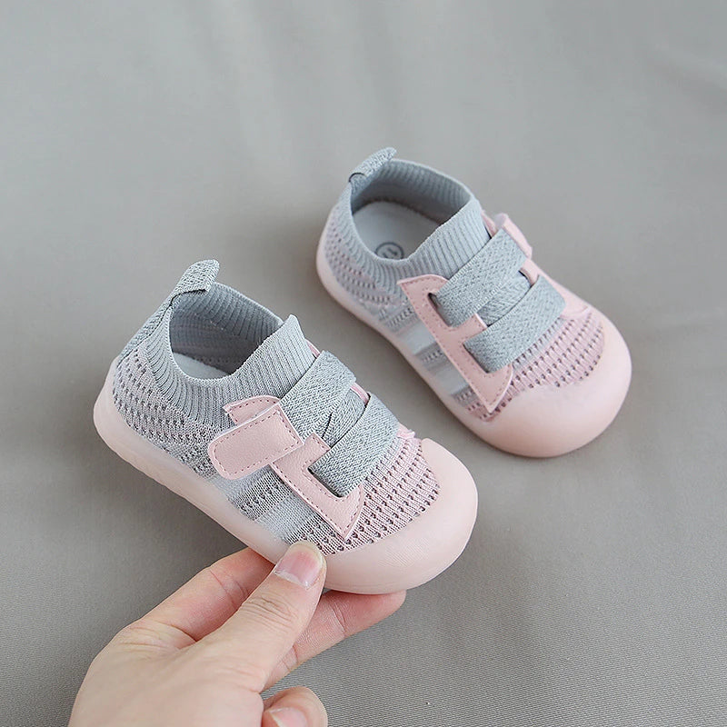 A pair of light pink and gray breathable mesh toddler walking shoes are shown being held against a neutral gray background.