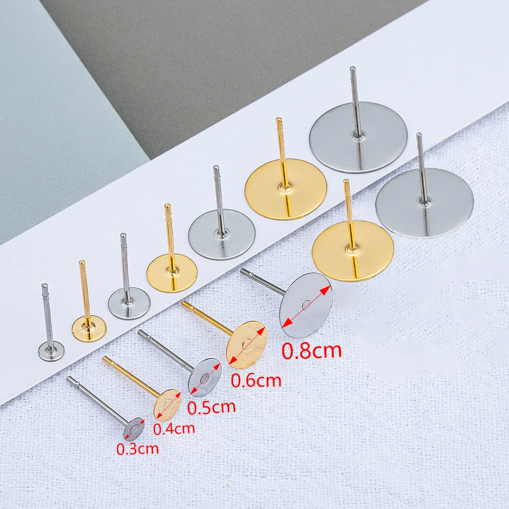 A collection of stainless steel earring stud bases in various sizes and gold and silver finishes laid out on a white surface.