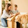 A woman is helping a boy use a cardboard construction tool set with a dog in a living room setting.