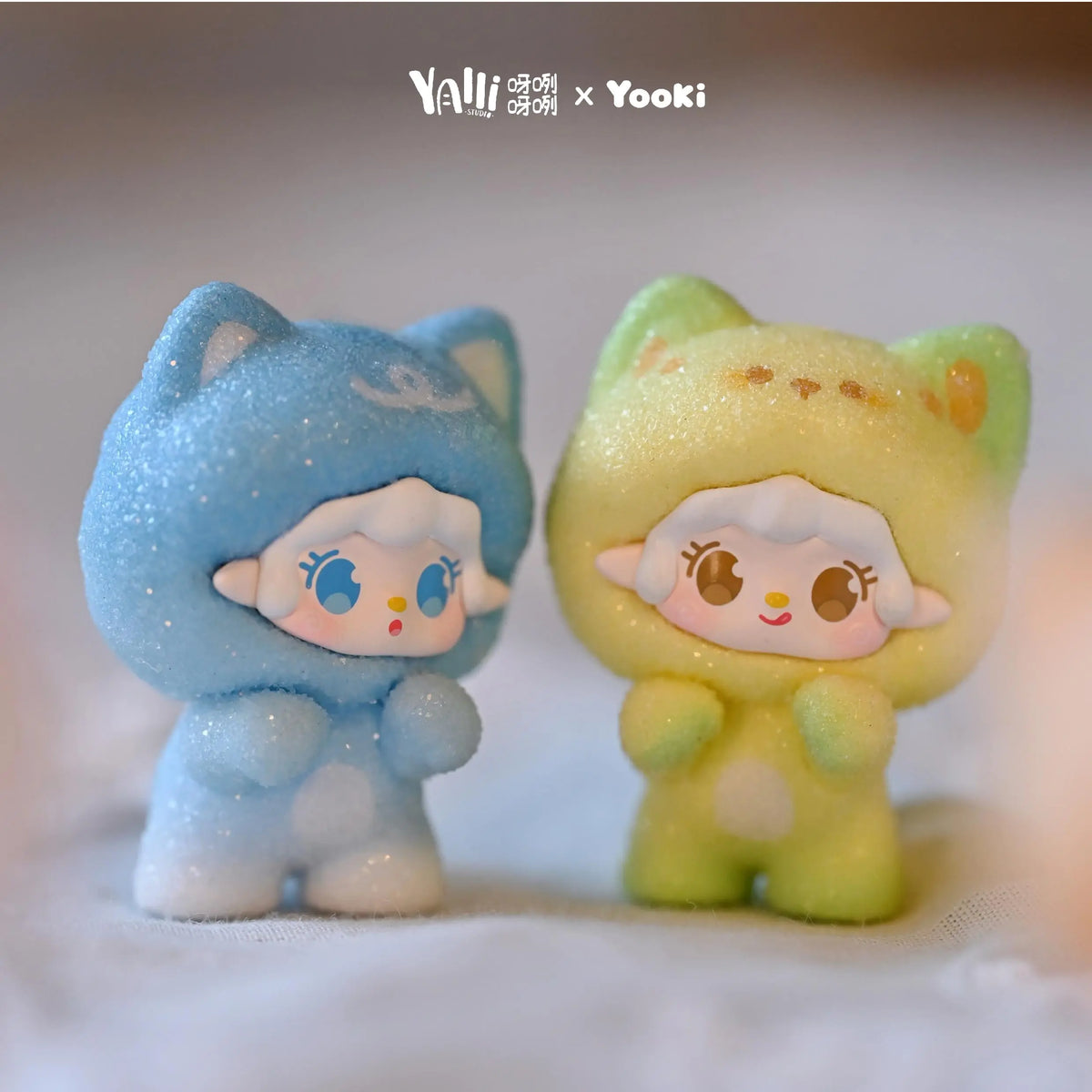A pair of cute, fuzzy cat figurines in blue and yellow are displayed together on a white surface.