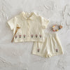 A cream baby cotton shirt and shorts set with a pink flower pattern laid on a white textured surface.