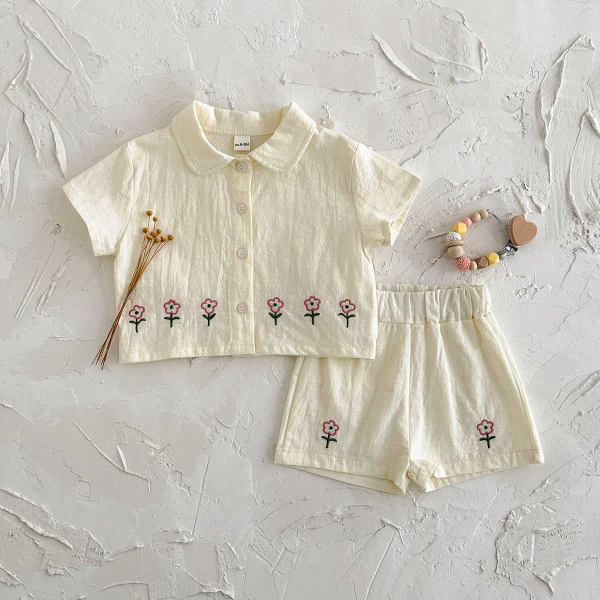 A cream baby cotton shirt and shorts set with a pink flower pattern laid on a white textured surface.
