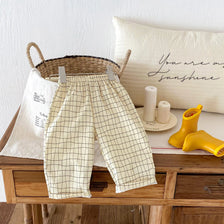 Khaki plaid casual pants with elastic waist displayed on a wooden table next to a pillow and yellow boots