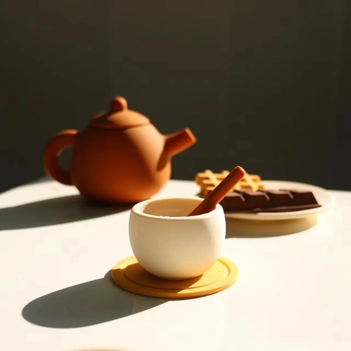 A small white cup sits on an orange saucer next to a brown teapot and a piece of chocolate on a white table.