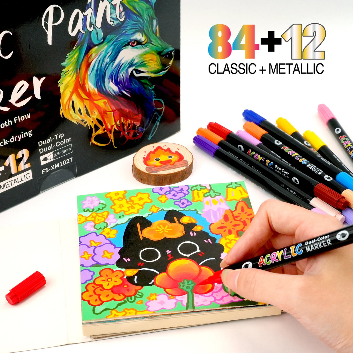 A hand holds a set of 12 classic and metallic dual-tip acrylic paint markers next to an open sketchbook.