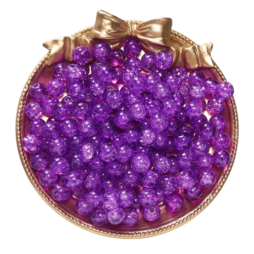 A pile of purple glass crackle beads for jewelry making displayed in a gold-toned dish with a bow.