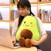 A young woman is holding a large, bright green avocado plush toy while working on a laptop in a bookshelf-lined room.