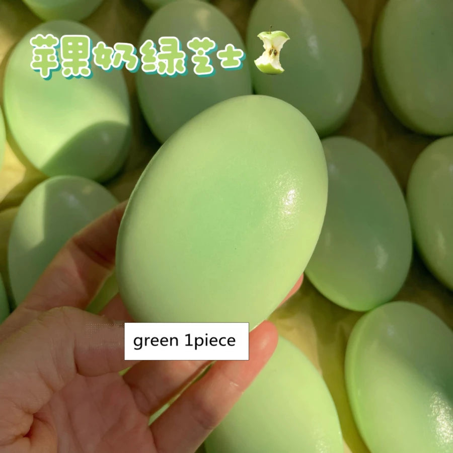A hand holds a green squishy cheese cake stress relief toy, showing one piece of the product.