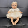 A lifelike 14-inch reborn baby doll with blonde hair and a white, ruffled fashion outfit sitting on a dark leather surface.