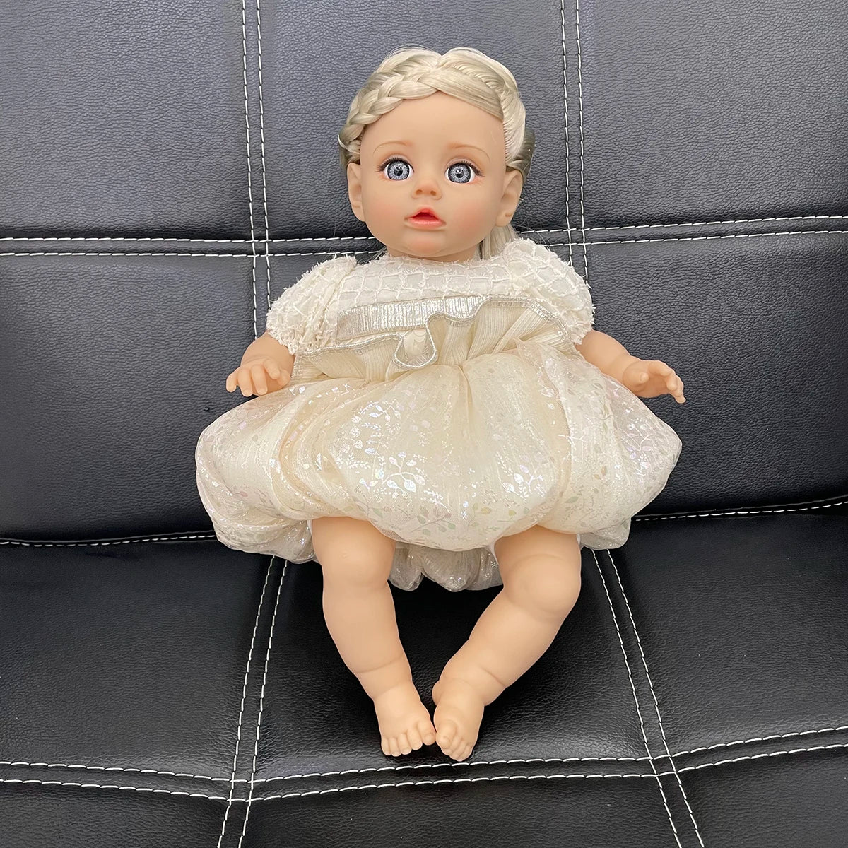 A lifelike 14-inch reborn baby doll with blonde hair and a white, ruffled fashion outfit sitting on a dark leather surface.