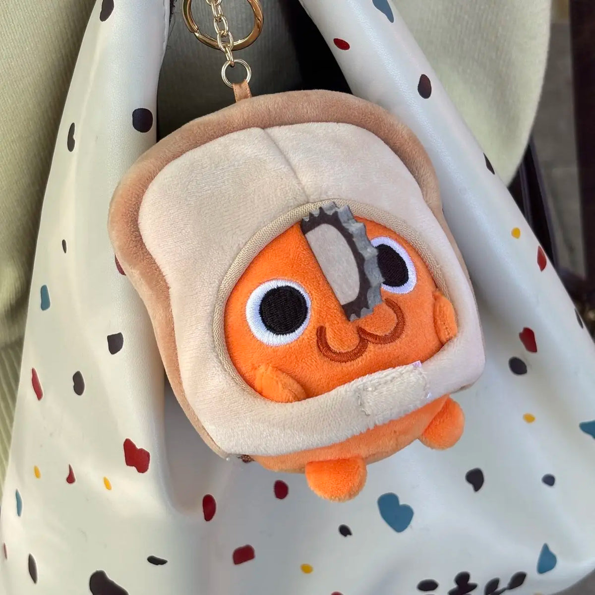 An orange Pochita plush doll with a chainsaw detail is hanging from a gold chain on a light-colored fabric bag.