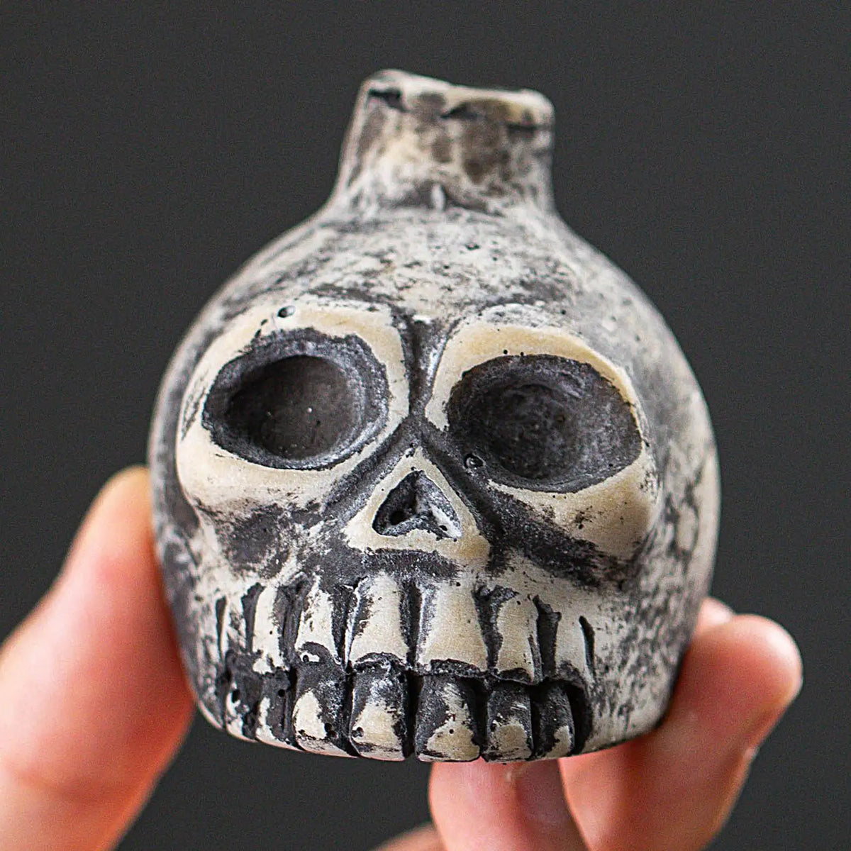A ceramic Aztec skull whistle with detailed facial features is held in a hand against a dark background.