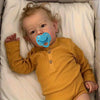 A baby wearing a yellow long-sleeved shirt and a blue pacifier is lying in a white bed.