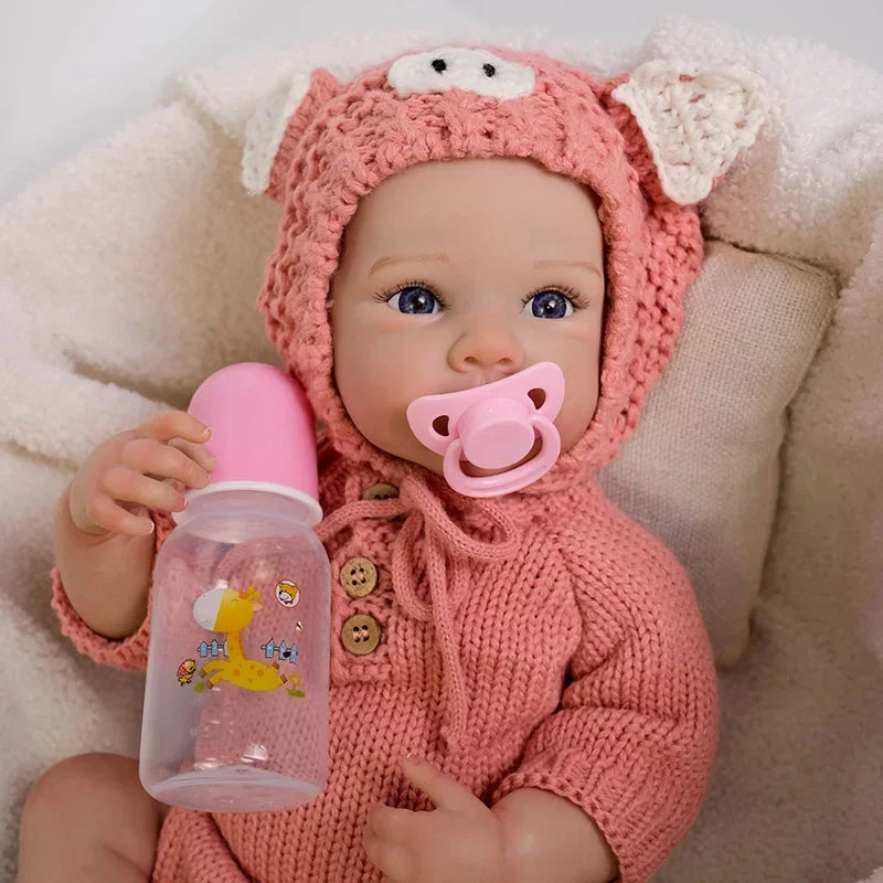 A realistic silicone reborn baby doll wearing a pink knitted animal hood and holding a pink bottle.