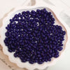 A pile of dark purple oval glass rice grain beads for jewelry making in a white dish.
