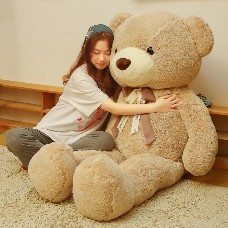 A young woman is sitting on a beige rug with a giant, light brown plush teddy bear.