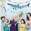 A group of children and an adult celebrating with a blue shark happy birthday banner and hanging garland set.