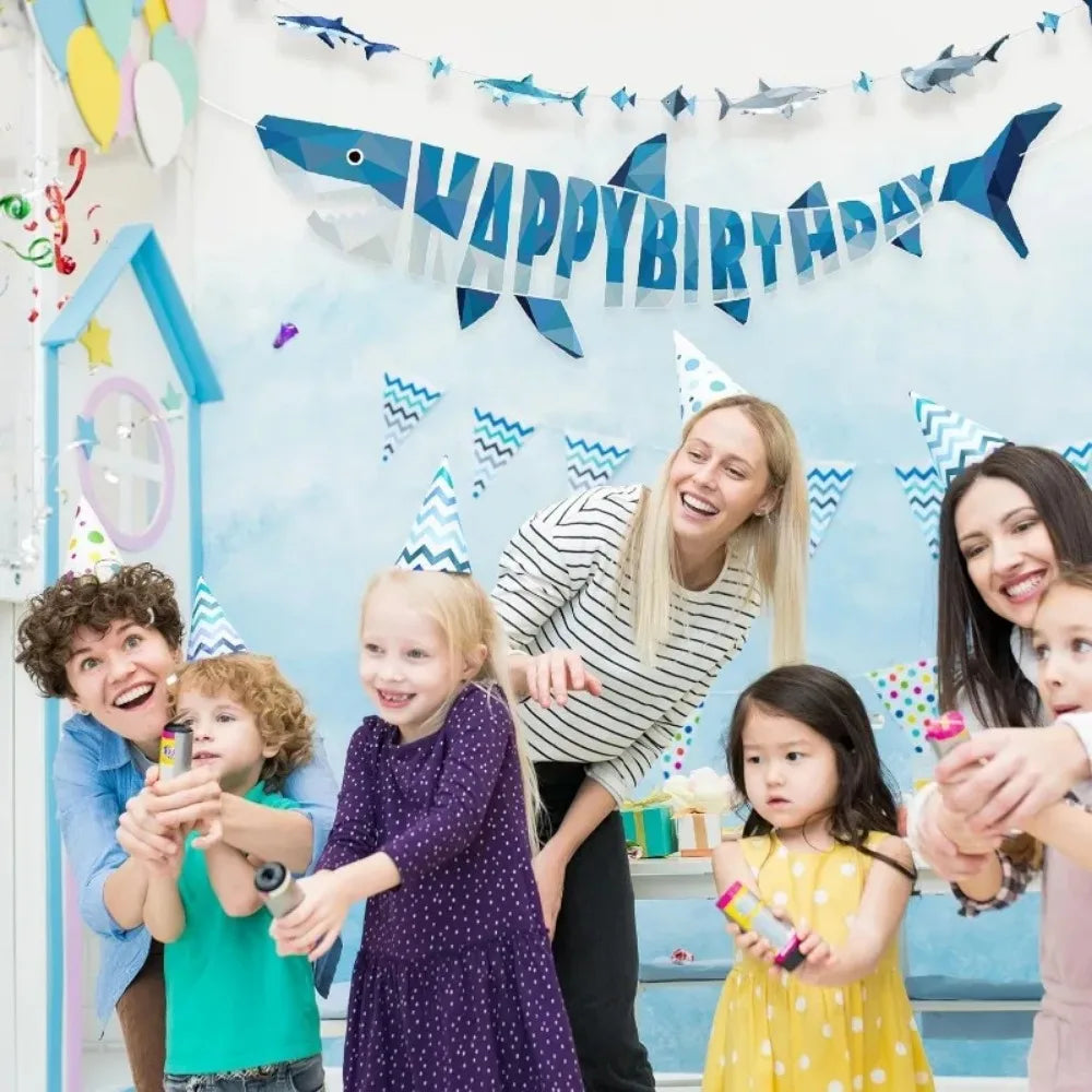 A group of children and an adult celebrating with a blue shark happy birthday banner and hanging garland set.