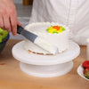 A hand uses a spatula to decorate a white frosted cake with orange and yellow floral decorations.