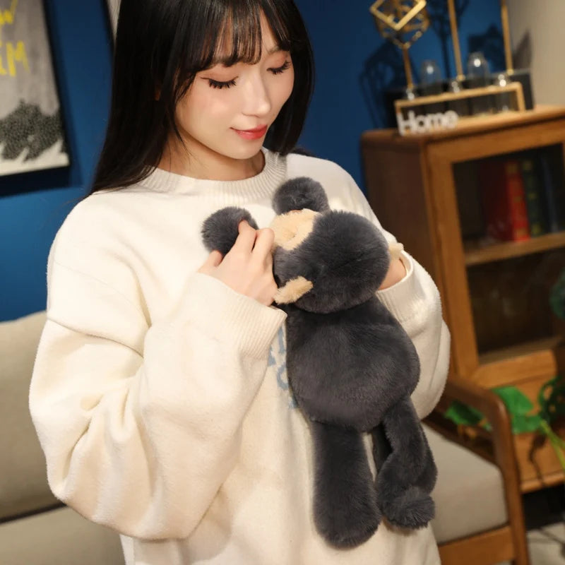A woman is holding a dark gray plush monkey soft toy against a white sweater.