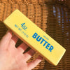 Eine Hand hält ein gelbes Squeeze-Butterprodukt mit 113g gesalzenem Butter auf einer geflochtenen Matte.