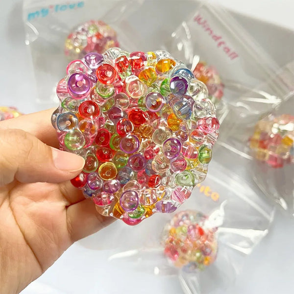 A hand holds a spherical cluster of colorful, translucent squishy beads in a clear plastic bag.