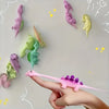 A hand holds a pink mini dinosaur finger slingshot, surrounded by various colorful plastic dinosaurs.