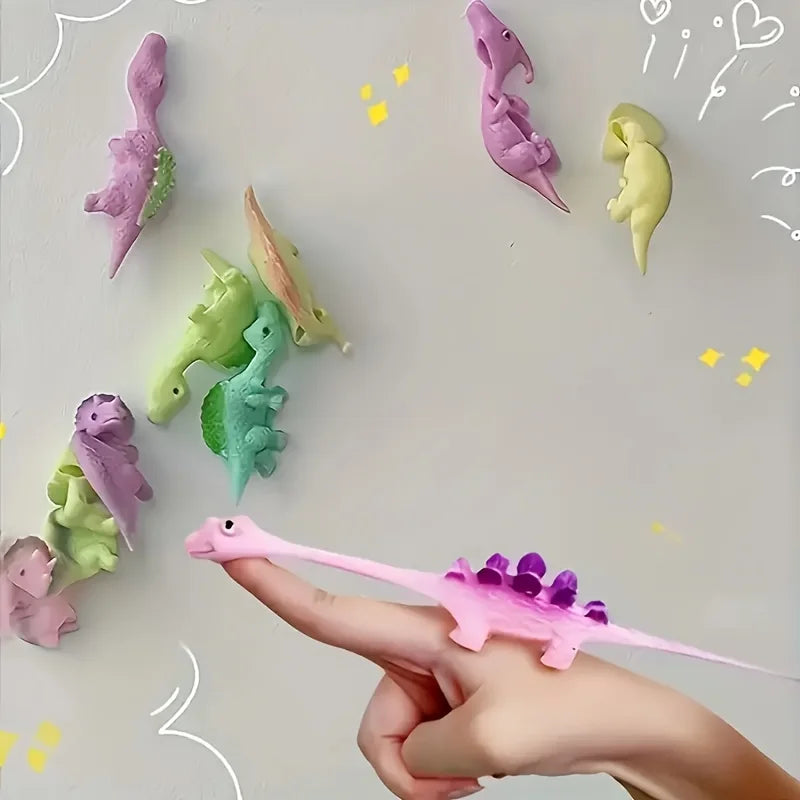 A hand holds a pink mini dinosaur finger slingshot, surrounded by various colorful plastic dinosaurs.