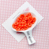 A white ceramic spoon holds bright orange plastic beads with a floral edge on a pink patterned background.