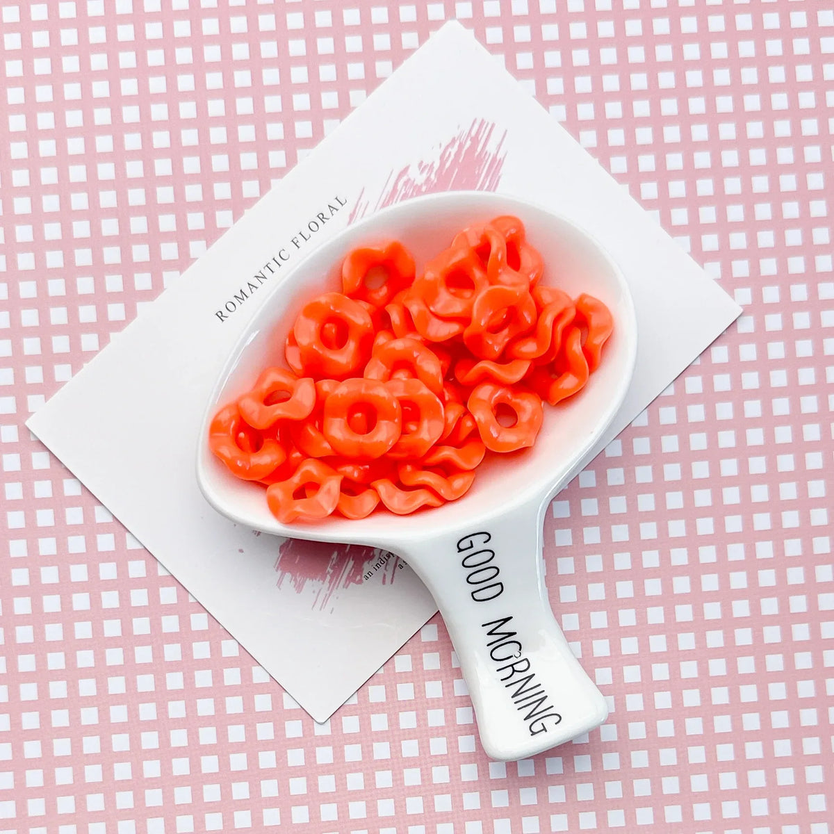 A white ceramic spoon holds bright orange plastic beads with a floral edge on a pink patterned background.
