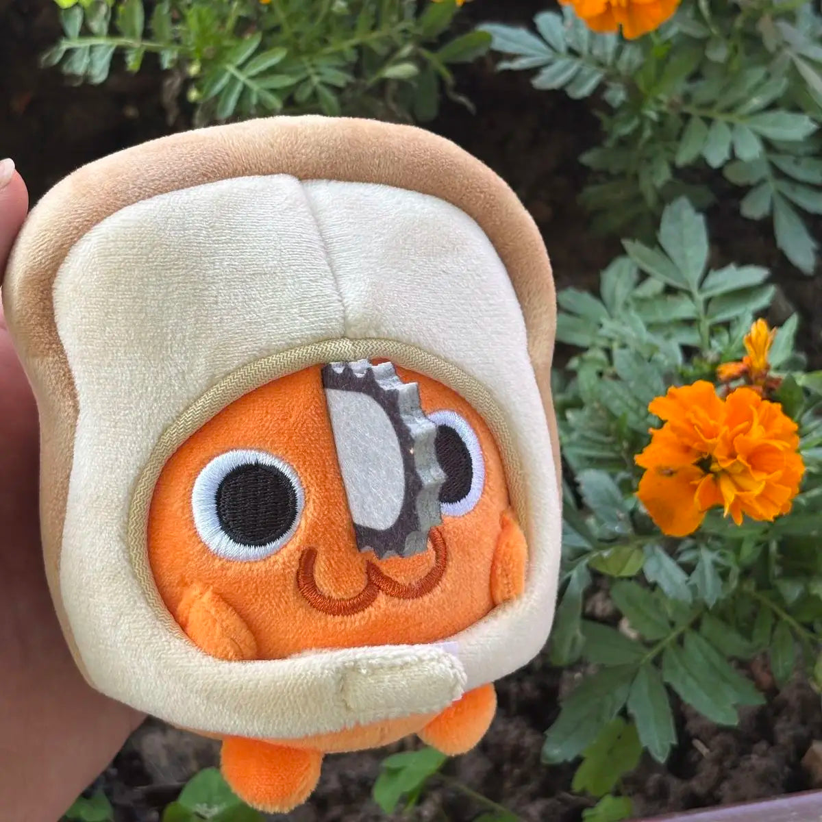 An orange Pochita plush doll with large eyes and a gray chainsaw accessory, set against green foliage.