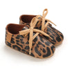 A pair of leopard print soft sole baby sneakers with tan laces and brown leather trim.
