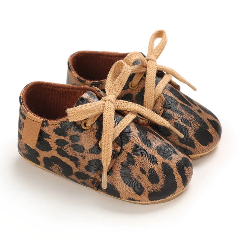 A pair of leopard print soft sole baby sneakers with tan laces and brown leather trim.