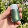 A hand holds a small glass bottle of metallic gold acrylic paint with a white cap against a blurred green outdoor background.