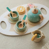 A wooden pretend play tea party set featuring a teapot, cups, and spoons in pastel green and natural wood tones.