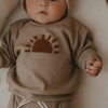 A baby wearing a beige long-sleeve outfit with a sun design on the chest.