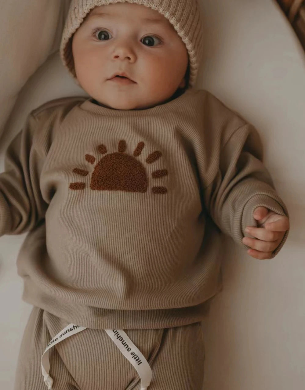 A baby wearing a beige long-sleeve outfit with a sun design on the chest.