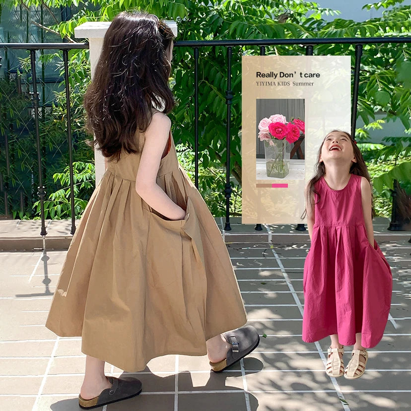 A young girl in a beige sleeveless cotton sundress stands outdoors with lush greenery in the background.